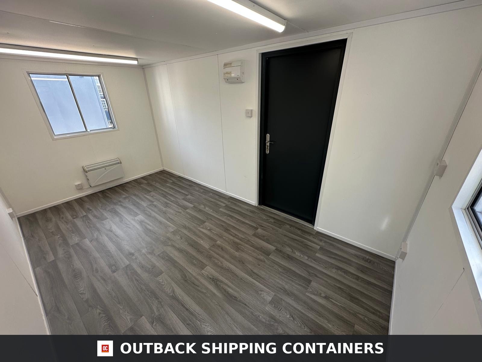 16x9 ft 16x9FT for Sale — Window and Shutter Detail | Outback Shipping Containers Interior view of a 16x9ft portable building with white walls, wood-effect flooring, a black door, and a wall heater