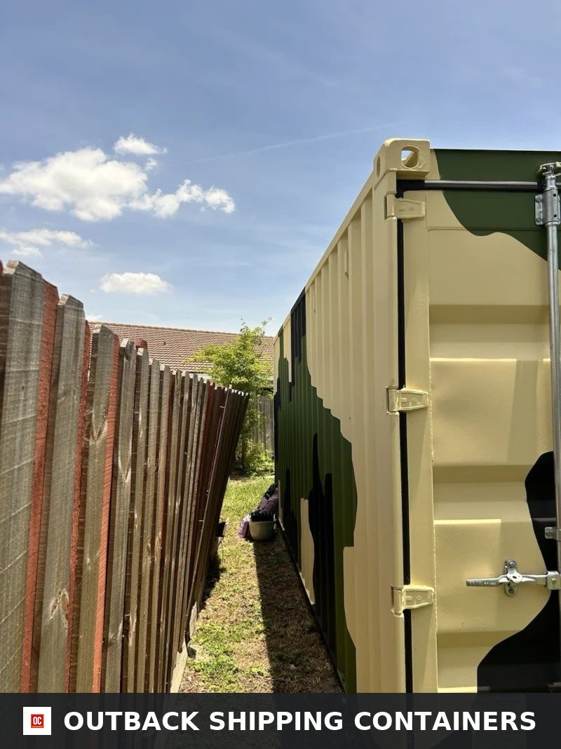 20ft Camouflage Storage Container — Three-Quarter Angle View | Outback Shipping Containers 20ft camouflage shipping container showing tight placement next to a wooden fence in a residential yard