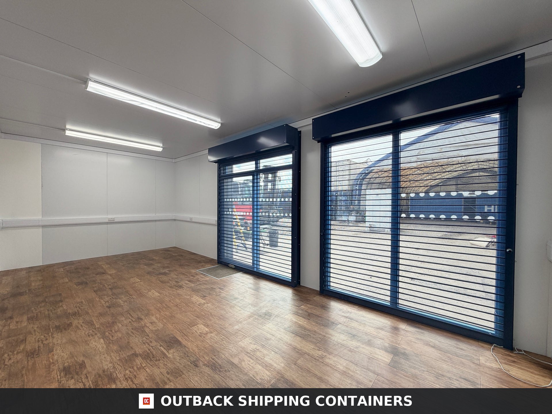 26x12ft for Sale — Interior Detail | Outback Shipping Containers Interior of a refurbished 26x12 marketing suite with wood style flooring, white walls, and blue shuttered glass doors