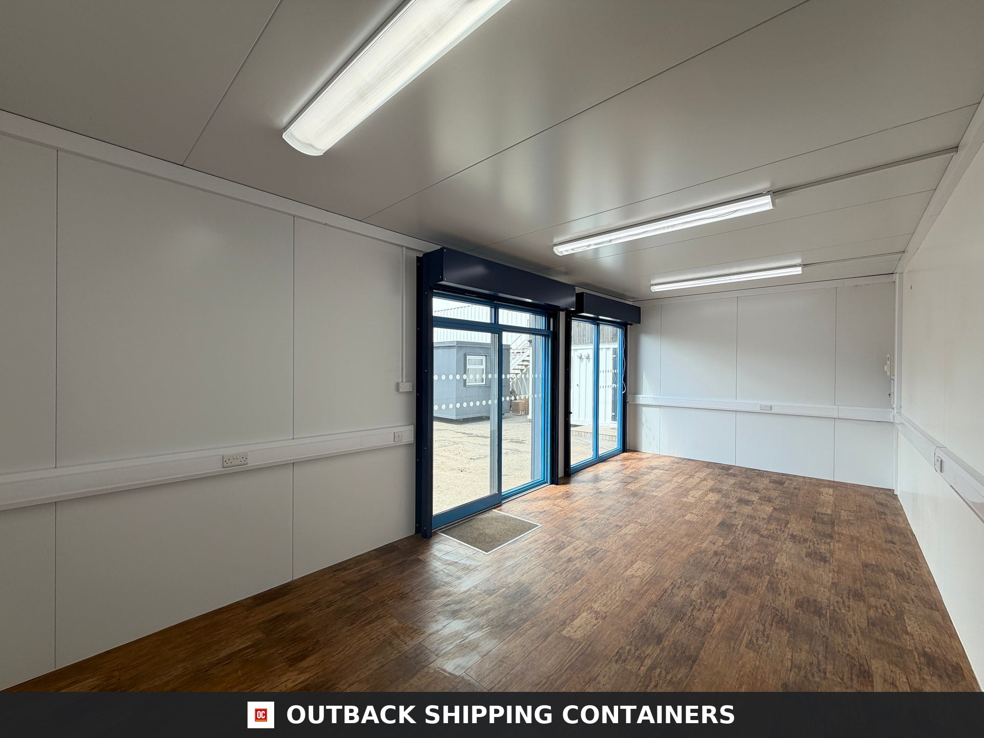 26x12ft for Sale — Rear Exterior View | Outback Shipping Containers Interior view of a 26x12ft portable marketing suite with wood flooring, white walls, and glass sliding doors