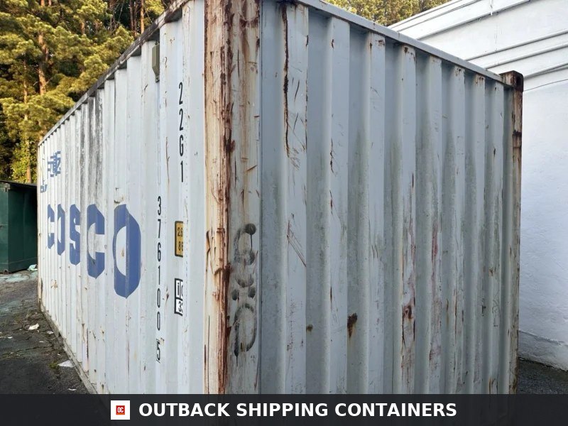 Side profile of a weathered grey 20ft shipping container with vertical ridges and surface rust parked outdoors
