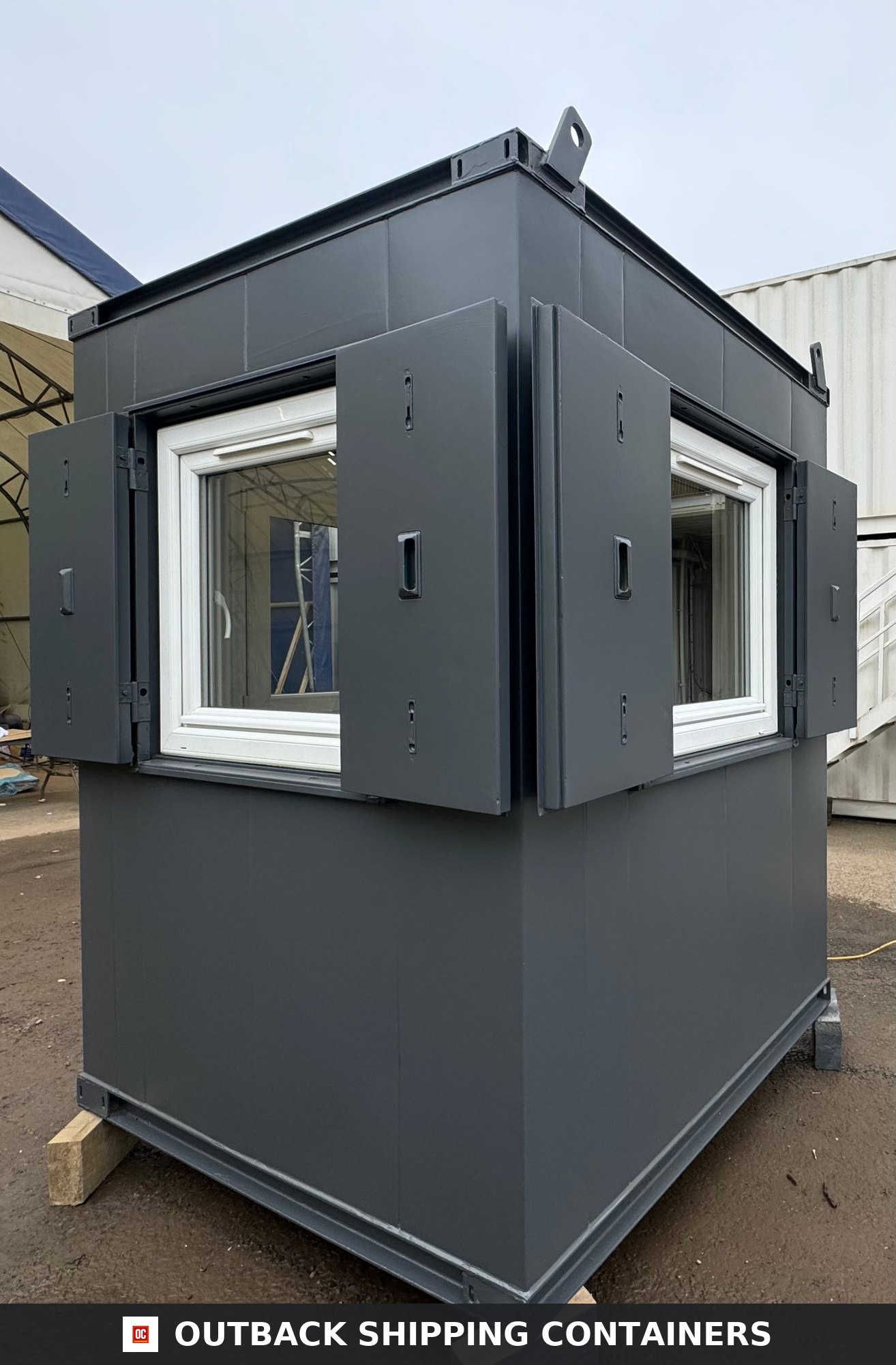 Dark grey 6x6 portable security hut with white window frames and open steel anti-vandal shutters on a flat lot