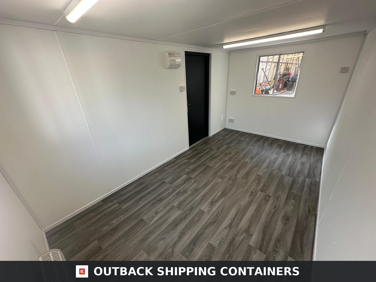16x9 ft 16x9FT for Sale — Rear Exterior View | Outback Shipping Containers Interior of a refurbished 16x9 ft portable building with white walls, wood-effect flooring, and overhead lighting