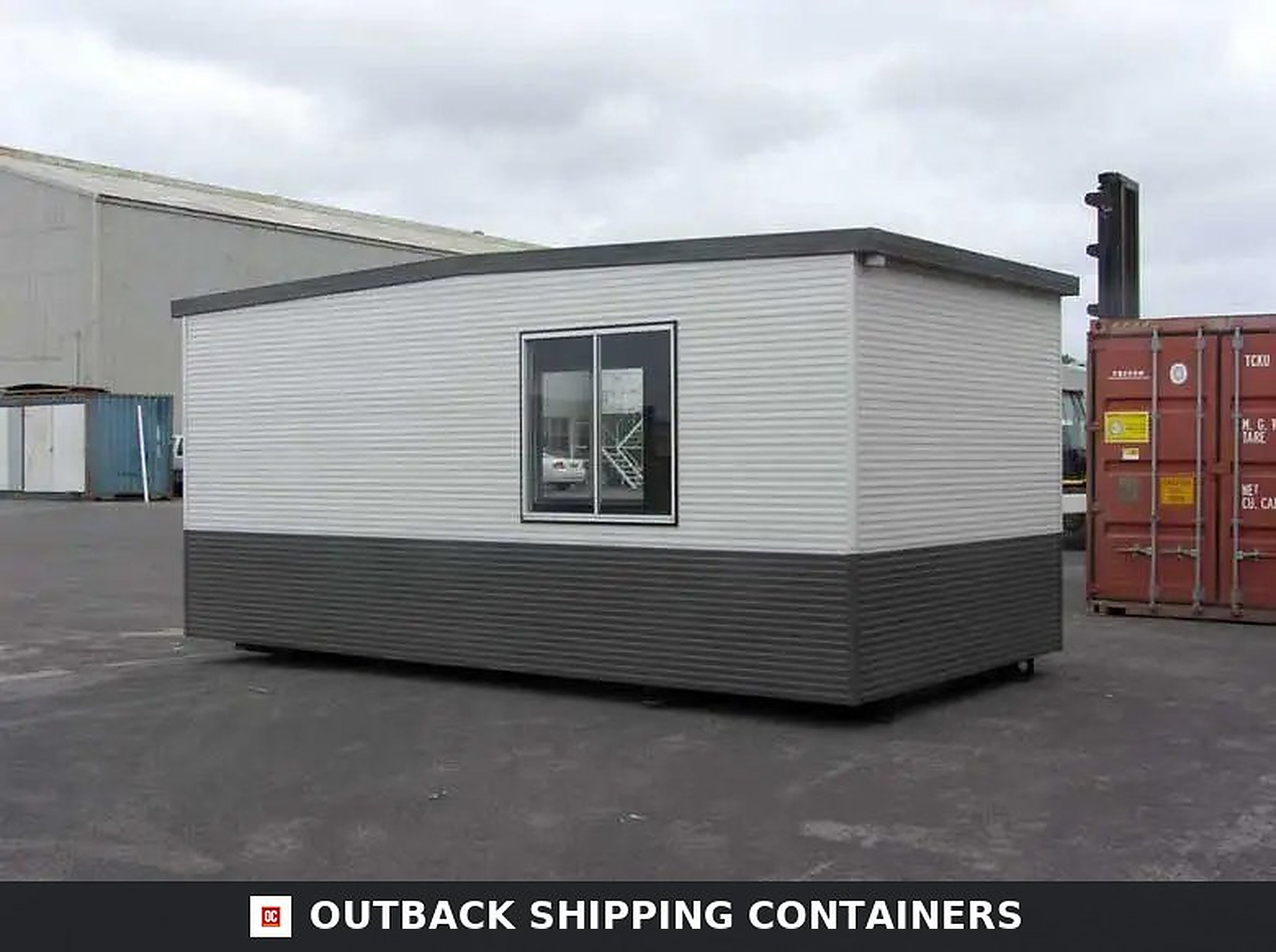 Modular container office with grey and white horizontal siding and a large central window on an asphalt lot