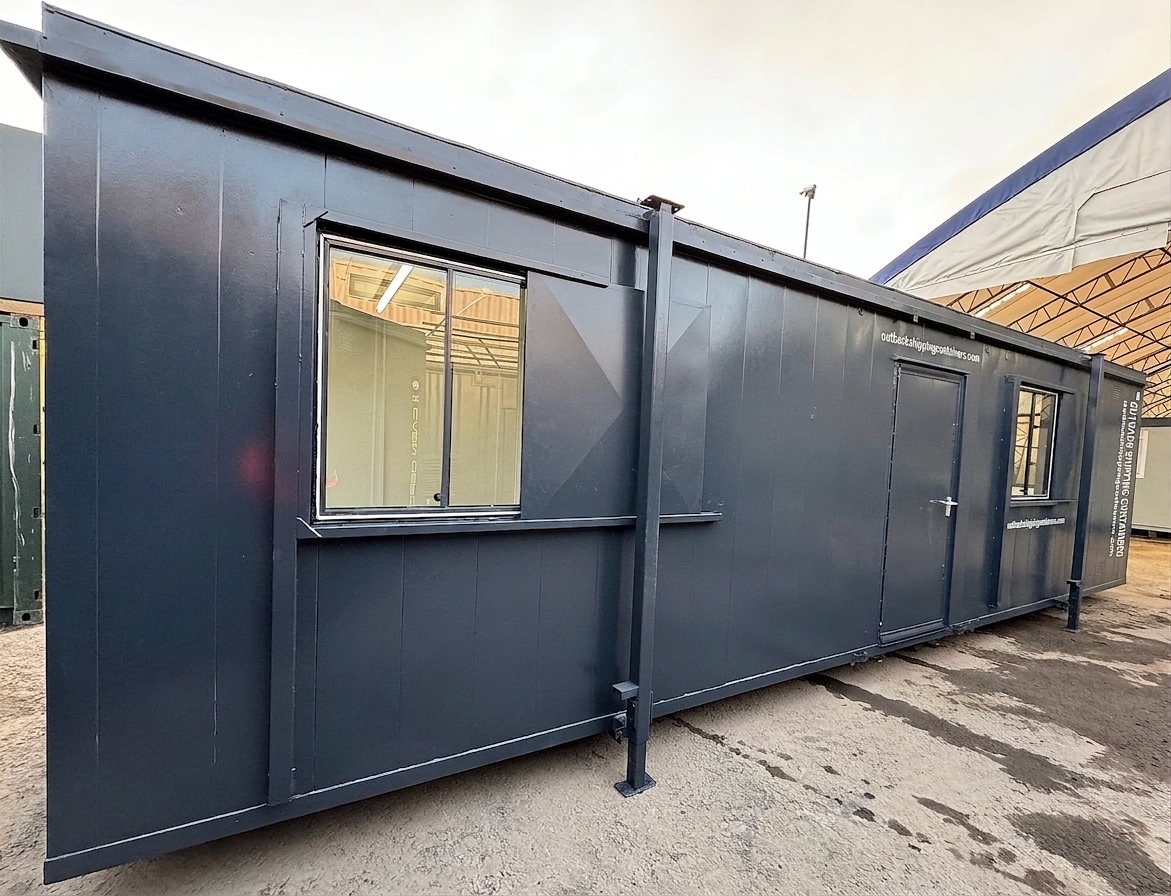 Side profile of a dark gray 32ft modular toilet facility with sliding glass windows and a secure steel door