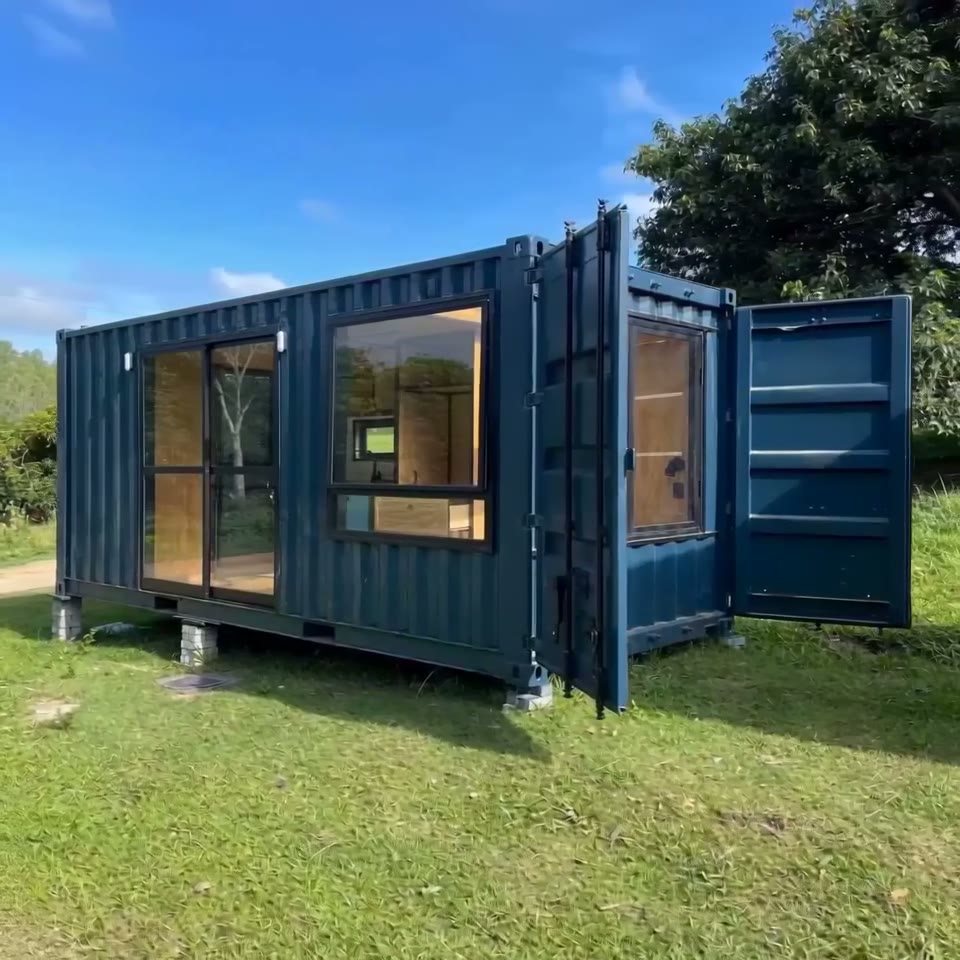 Exterior view of a dark blue 20ft shipping container cabin with large glass sliding doors and windows on a grassy field