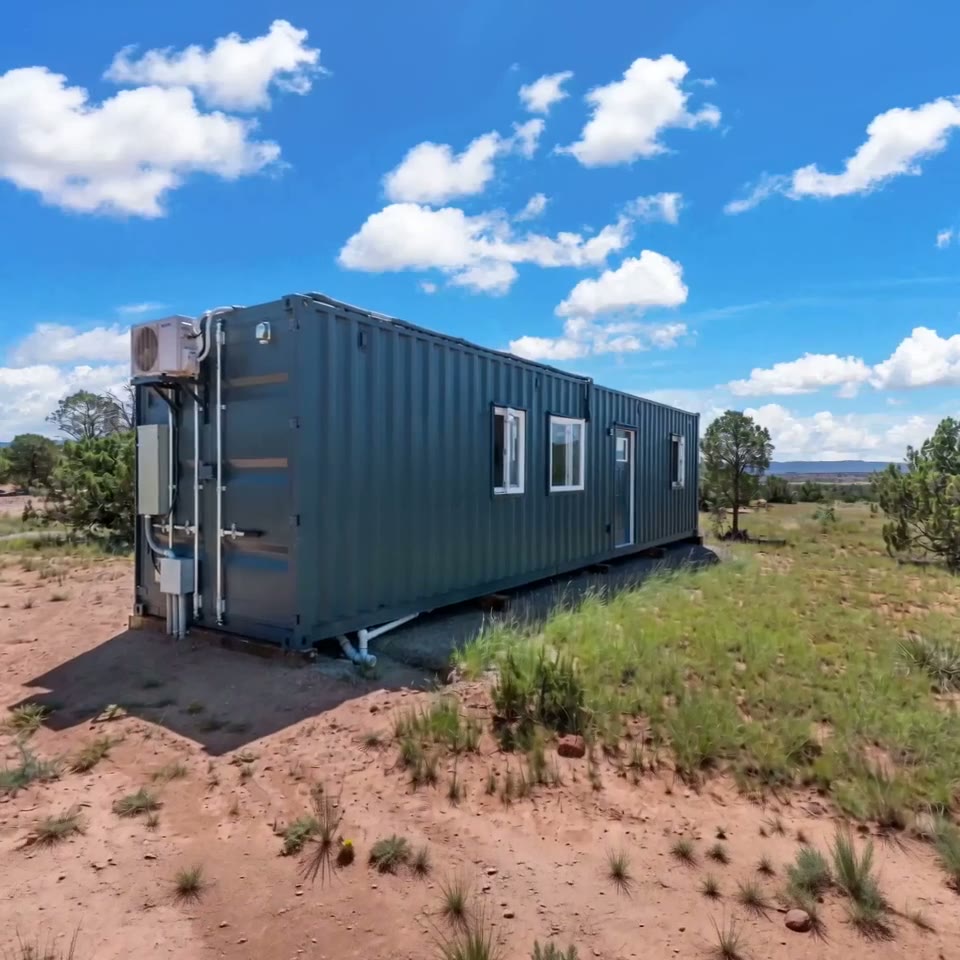 Exterior view of a dark gray 20ft shipping container home with multiple windows and an HVAC unit under a blue sky