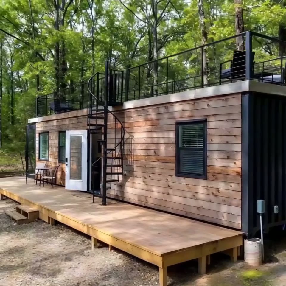 Exterior of a 40ft container home featuring wood siding, a spiral staircase, and a rooftop deck in a forest setting