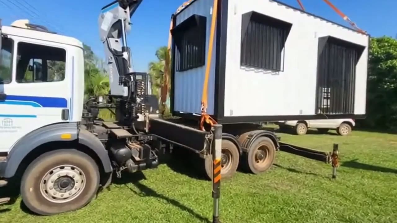 A white 20ft shipping container home with black window shutters being lifted by a crane truck